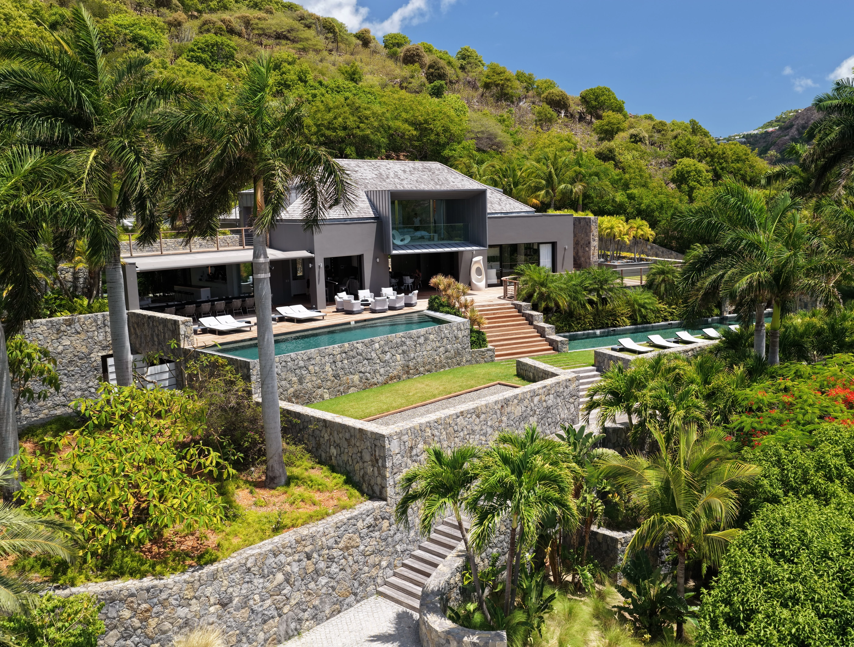 The house from above in daylight: the pools and the loungers on the terraces, the stone walls stepping down through the palms, the hills behind